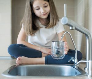 A child sitting cross-legged on the counter, filling up a glass cup from the tap.